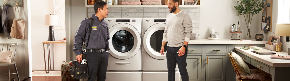 A laundry room where a woman is loading clothes into a Whirlpool Front-Load Washer that's next to a Whirlpool Front-Load Dryer. Also in the room are a desk and chair, a plant in a vase and various items on the counters. Through the doorway is a small table with a lamp and a potted plant