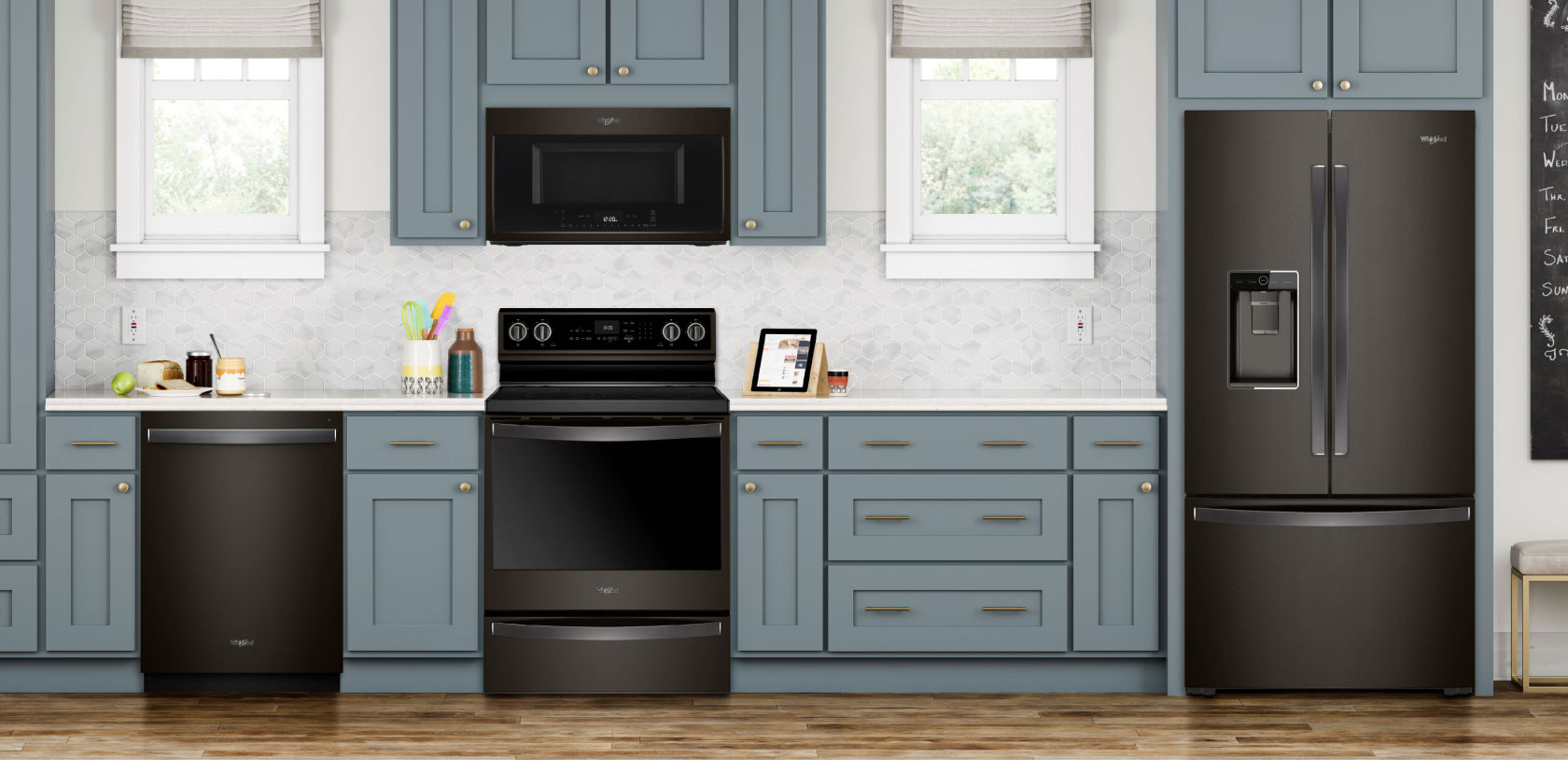 A kitchen with an island and three stools. On the island is a bowl of fruit on a placemat. The cupboards are grey and the Whirlpool Black Stainless Steel Appliances include a refrigerator, an oven with an over-the-range microwave and a dishwasher. On the counters are a kitchen utensil holder, cutting boards, a potted plant, salt and pepper shakers and more