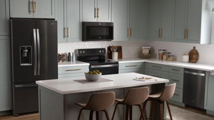 A kitchen with an island and three stools. On the island is a bowl of fruit on a placemat. The cupboards are grey and the Whirlpool Black Stainless Steel Appliances include a refrigerator, an oven with an over-the-range microwave and a dishwasher. On the counters are a kitchen utensil holder, cutting boards, a potted plant, salt and pepper shakers and more