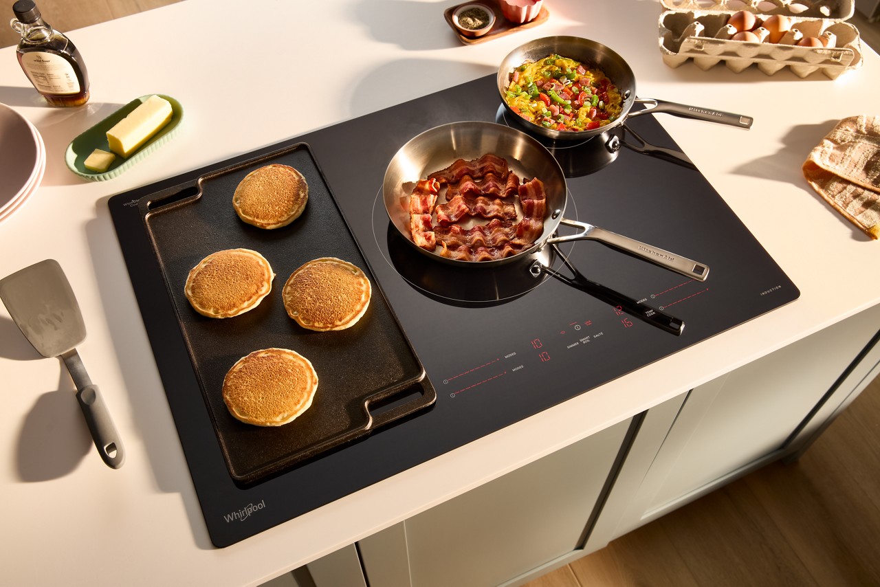 A Whirlpool Cooktop with a griddle. On the griddle are four pancakes. On the burners are two pots with ingredients. Various items, like butter in a butter dish, are on the counter.