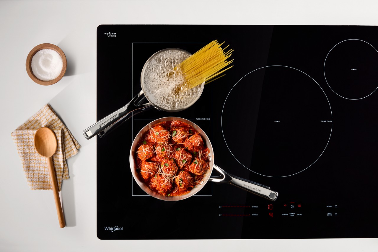 On a Whirlpool Cooktop, meatballs cook in a pot and pasta boils in another. Next to the cooktop is a wooden spoon on a dishcloth and a small bowl of salt. 