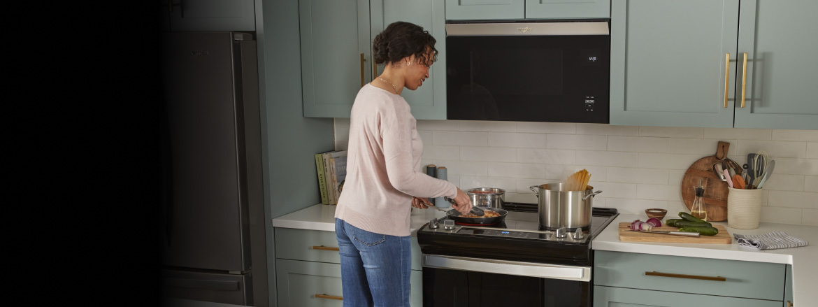 Une femme cuisine avec une poêle sur une cuisinière Whirlpool située sous un four à micro-ondes encastré. Il y a également deux casseroles sur la cuisinière, dont l'une contient des pâtes en train de cuire dans de l'eau bouillante. Il y a aussi un réfrigérateur Whirlpool. Sur le plan de travail, il y a des livres de cuisine, une planche à découper avec un oignon rouge, des concombres et un couteau, un torchon, un porte-ustensiles de cuisine et une autre planche à découper appuyée contre le mur