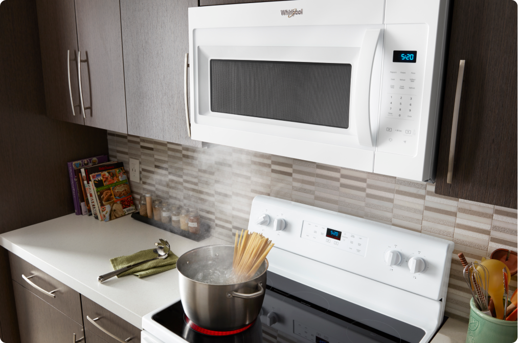 Des pâtes cuisent sur une cuisinière Whirlpool sous un four à micro-ondes à hotte intégrée Whirlpool. Sur les comptoirs se trouvent une louche posée sur un linge à vaisselle, des livres de cuisine et un porte-ustensiles