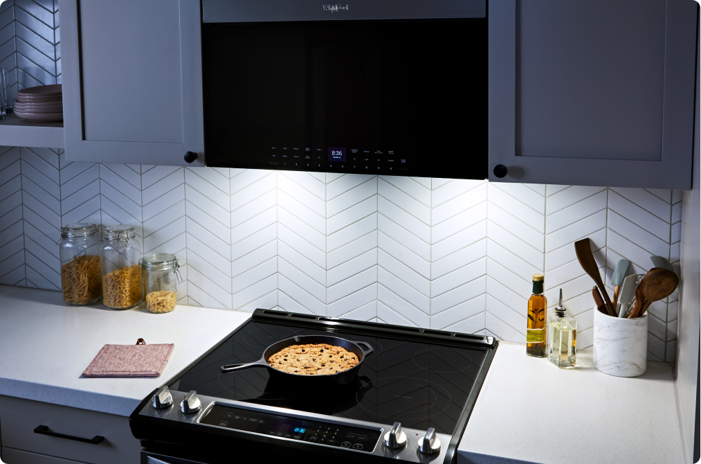 Un four à micro-ondes à hotte intégrée Whirlpool au un design affleurant. Sa lumière éclaire la table de cuisson et une casserole en dessous. Sur le comptoir se trouvent un porte-ustensiles, des contenants d'huile et des bocaux de pâtes