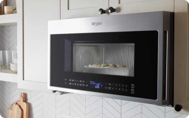 Un four à micro-ondes Whirlpool dans lequel cuisent des aliments. À proximité se trouvent des planches à découper et des étagères contenant des objets comme des verres