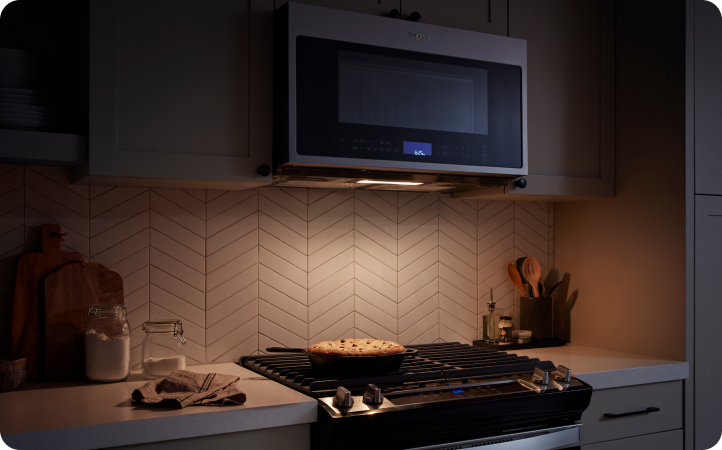 In a dark kitchen is a Whirlpool Microwave over a Whirlpool Cooktop. The cooktop light is on and a pan of food cooks. On the counters are cutting boards, a utensil holder and other items