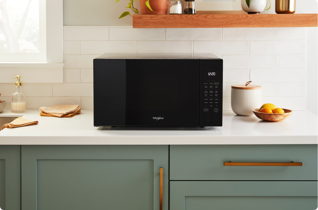 A Whirlpool Countertop Microwave next to a bowl of fruit, a container and dishcloths. Above is a shelf with a plant and other items