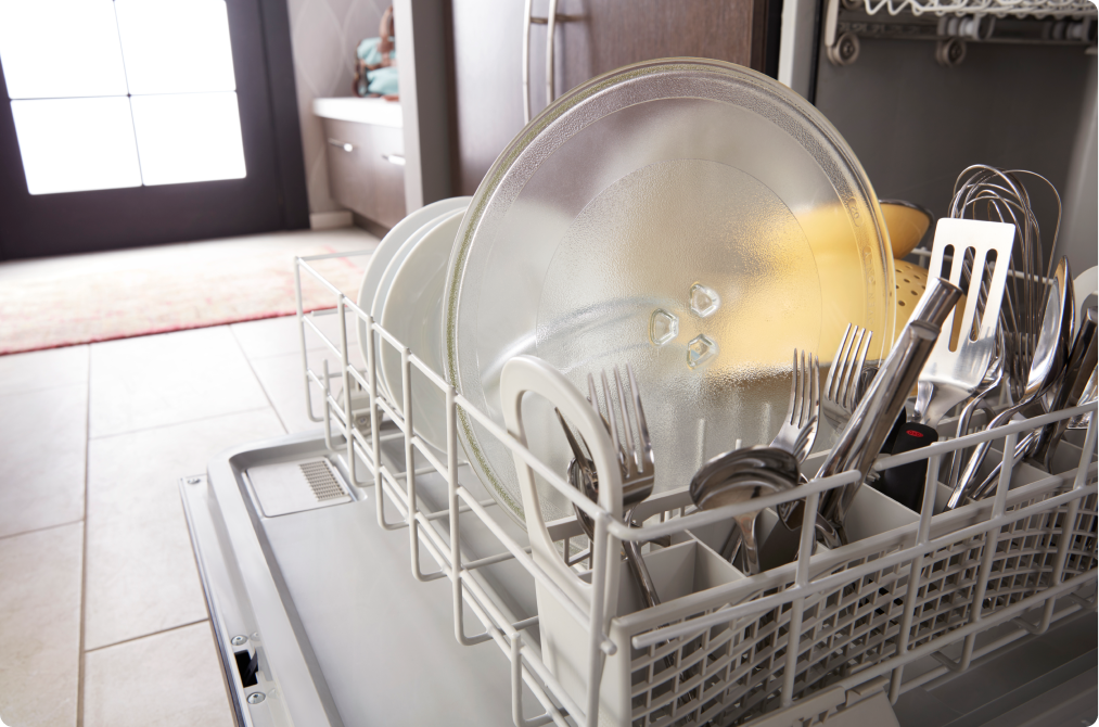 Un lave-vaisselle Whirlpool ouvert avec le panier sorti, dans lequel se trouvent des ustensiles, un plateau tournant, des des assiettes et une passoire