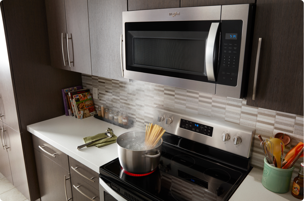 A kitchen with a Whirlpool Over-the-Range Microwave above a Whirlpool Range. On the induction cooktop, pasta boils in a pot of water. On the counters are a utensil holder, a utensil on a cloth and cookbooks