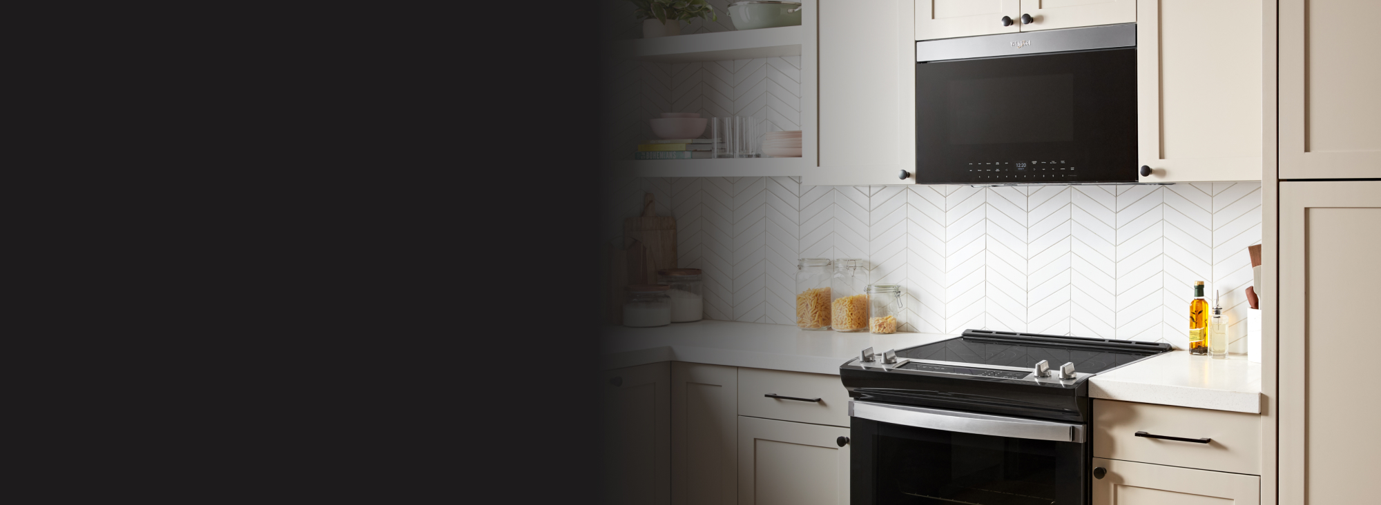A white kitchen with a Whirlpool Over-the-Range Microwave with a flush design over a Whirlpool Induction Range. On the counters are glass jars of pasta and flour, oil containers and cutting boards. On the above shelves are cookbooks, glasses and bowls