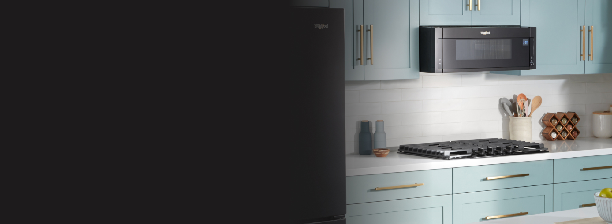 Une cuisine avec des armoires vertes est équipée d'un four à micro-ondes Whirlpool au-dessus d'une table de cuisson Whirlpool. Sur les comptoirs se trouvent une salière et une poivrière, un porte-ustensiles, une étagère à épices, un petit bol en bois et un récipient