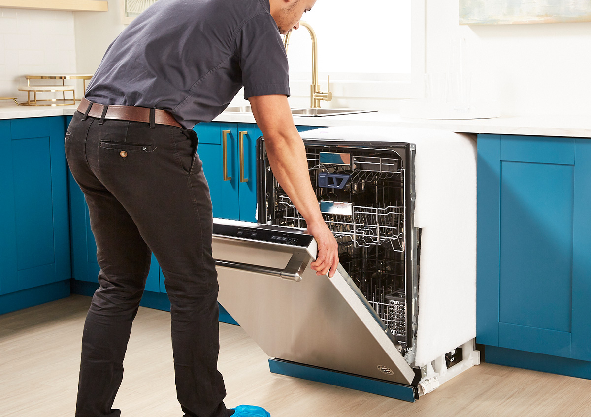 A uniformed repairman installs a dishwasher in a kitchen with blue cabinets
