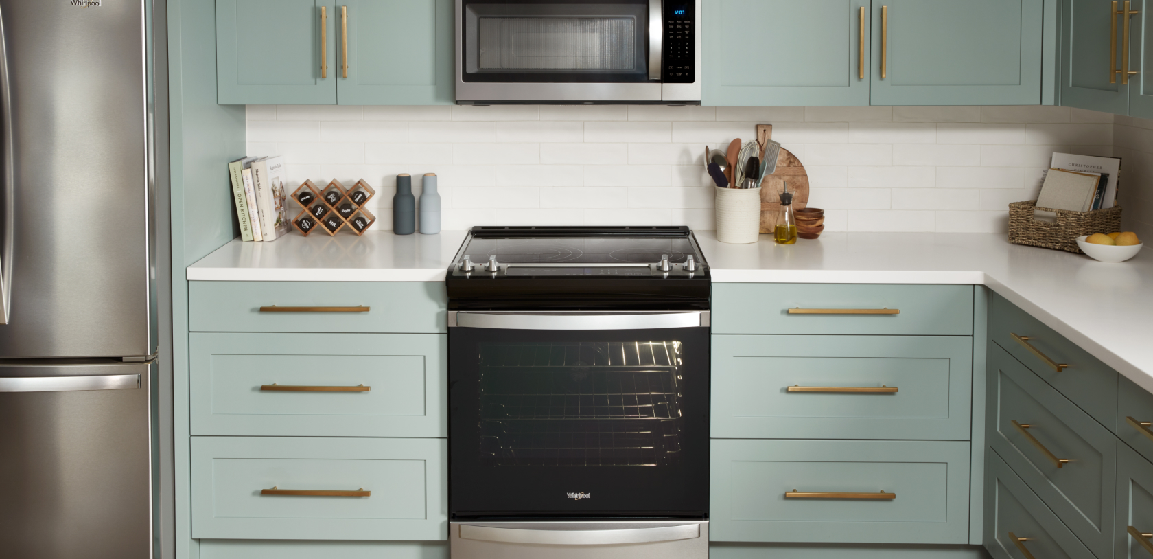 A kitchen with a Whirlpool Refrigerator and a Whirlpool Range with an Over-the-Range Microwave. On the counters are cookbooks, a spice rack, a full kitchen utensil holder, a cutting board and more