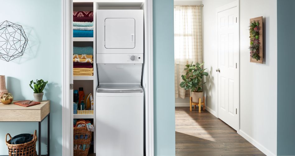 A room with a Whirlpool stacked washer and dryer combination unit. Next to it are shelves with towels and supplies. Under the shelves is a basket with laundry. Next to the laundry area is a table with a potted plant on top and a basket underneath.