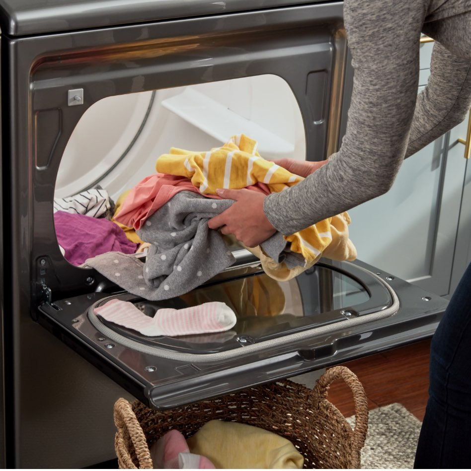 The hamper of a dryer is opened. Someone wearing a grey shirt is pulling out grey, pink and yellow laundry.
