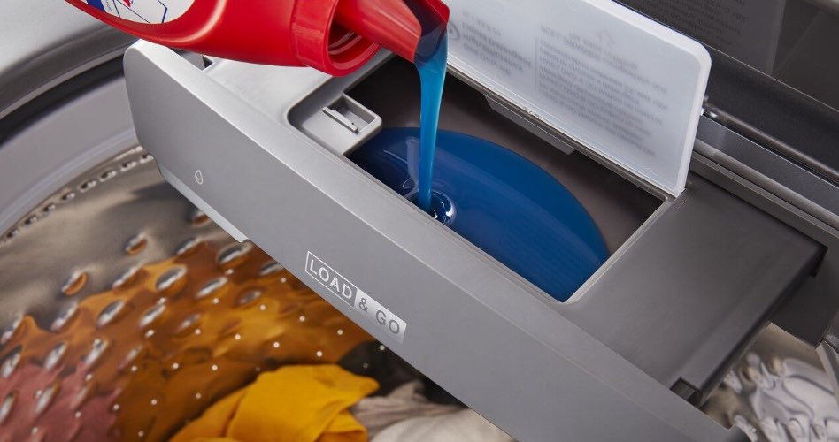 Someone pouring blue liquid laundry detergent from a red and white bottle into the dispenser of a washing machine.