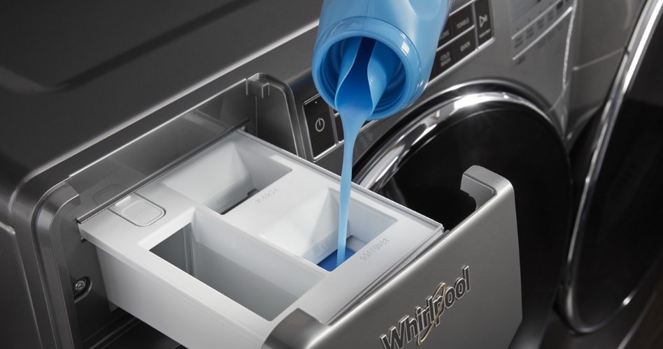blue liquid laundry detergent being poured out of the spout of a bottle into the dispenser of a washing machine.