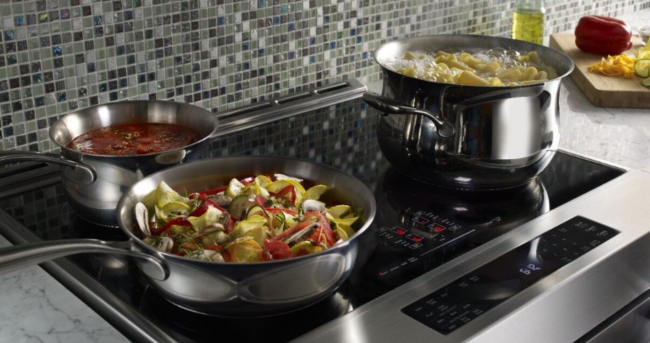 ne cuisinière à induction Whirlpool avec un chaudron de pâtes dans l'eau qui bout, une casserole remplie de légumes et une autre de sauce. Sur le comptoir, une bouteille d'huile et une planche à découper avec un poivron rouge, des concombres en tranches et un poivron jaune en dés.