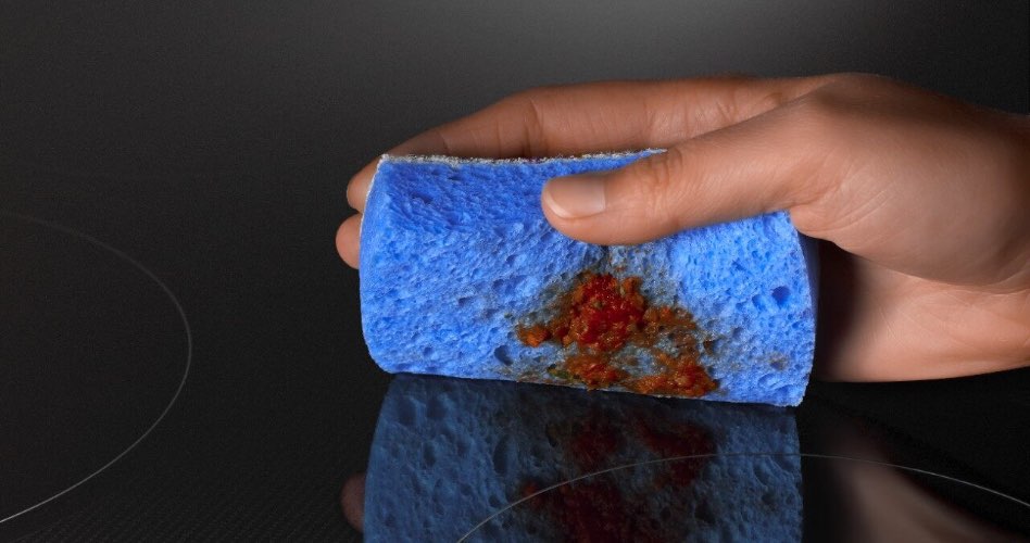 Quelqu'un nettoie une table de cuisson à induction avec une éponge bleue. Sur l'éponge se trouve de la sauce rouge.