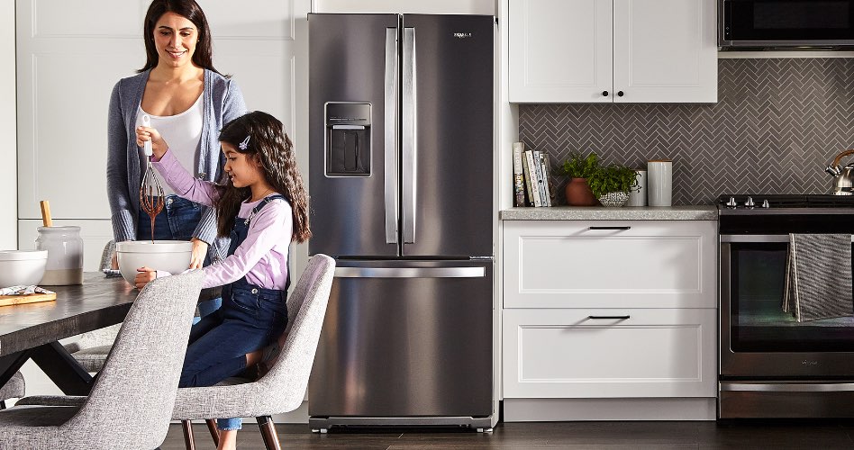 Une femme et un enfant à la table de cuisine. L'enfant soulève d'un bol un batteur couvert de chocolat. En arrière-plan, un réfrigérateur à portes françaises KitchenAid.