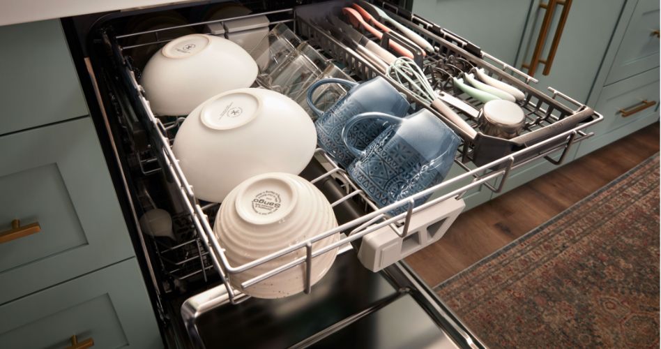 An opened Whirlpool dishwasher with the top rack pulled out. On the rack are bowls, mugs, glasses, cutlery and utensils.
