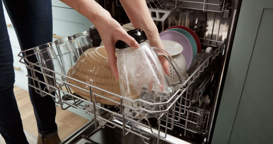 Someone loads a blender jar onto the middle rack of a Whirlpool Dishwasher. Also on the rack are glasses, bowls and plates.