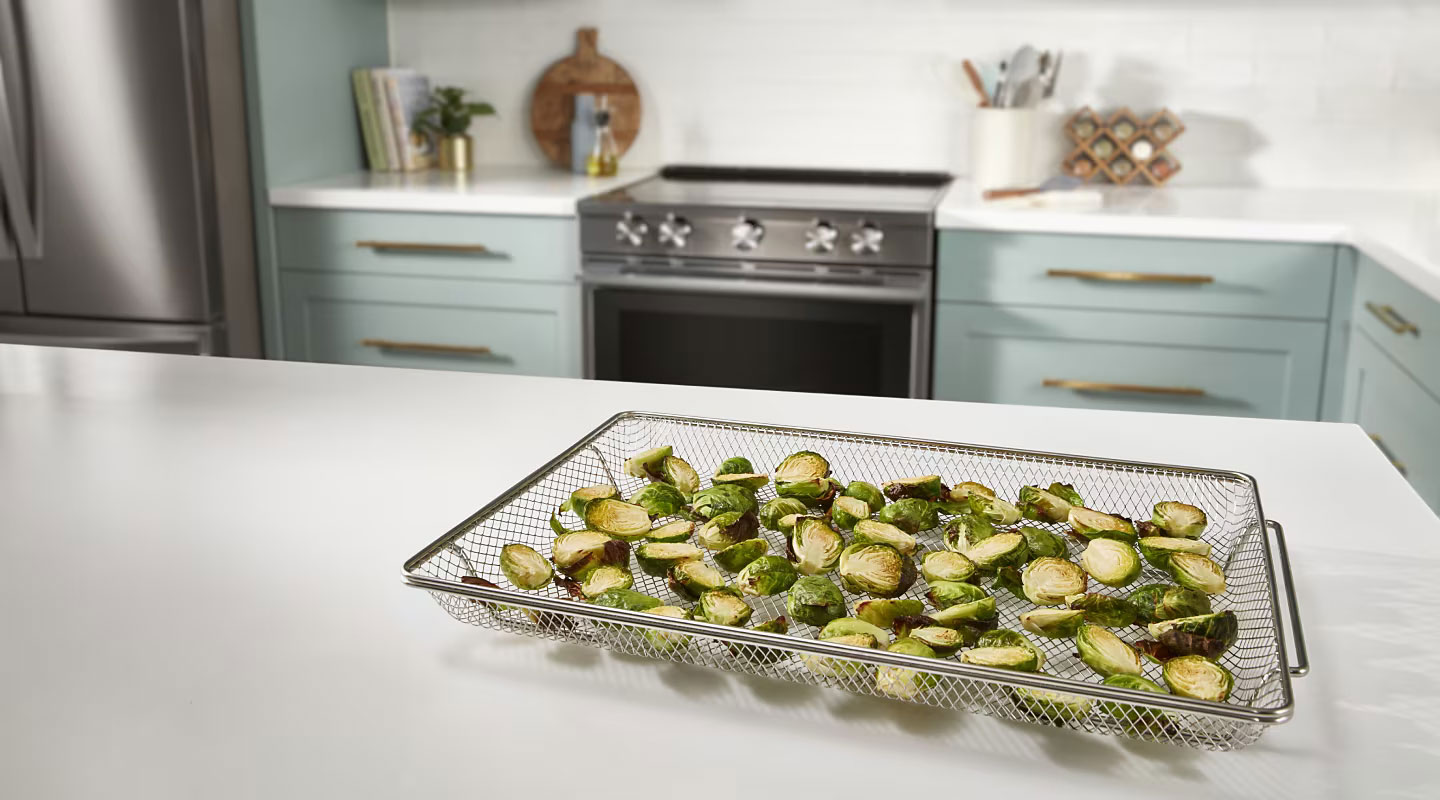Whirlpool Combination Microwave Une cuisine avec un réfrigérateur et une cuisinière Whirlpool. Sur l'îlot se trouve un panier de friture à air avec des tranches de courgettes. Sur les comptoirs à l'arrière-plan se trouvent des livres de cuisine, une planche à découper, une étagère à épices et un porte-ustensiles de cuisine, entre autres