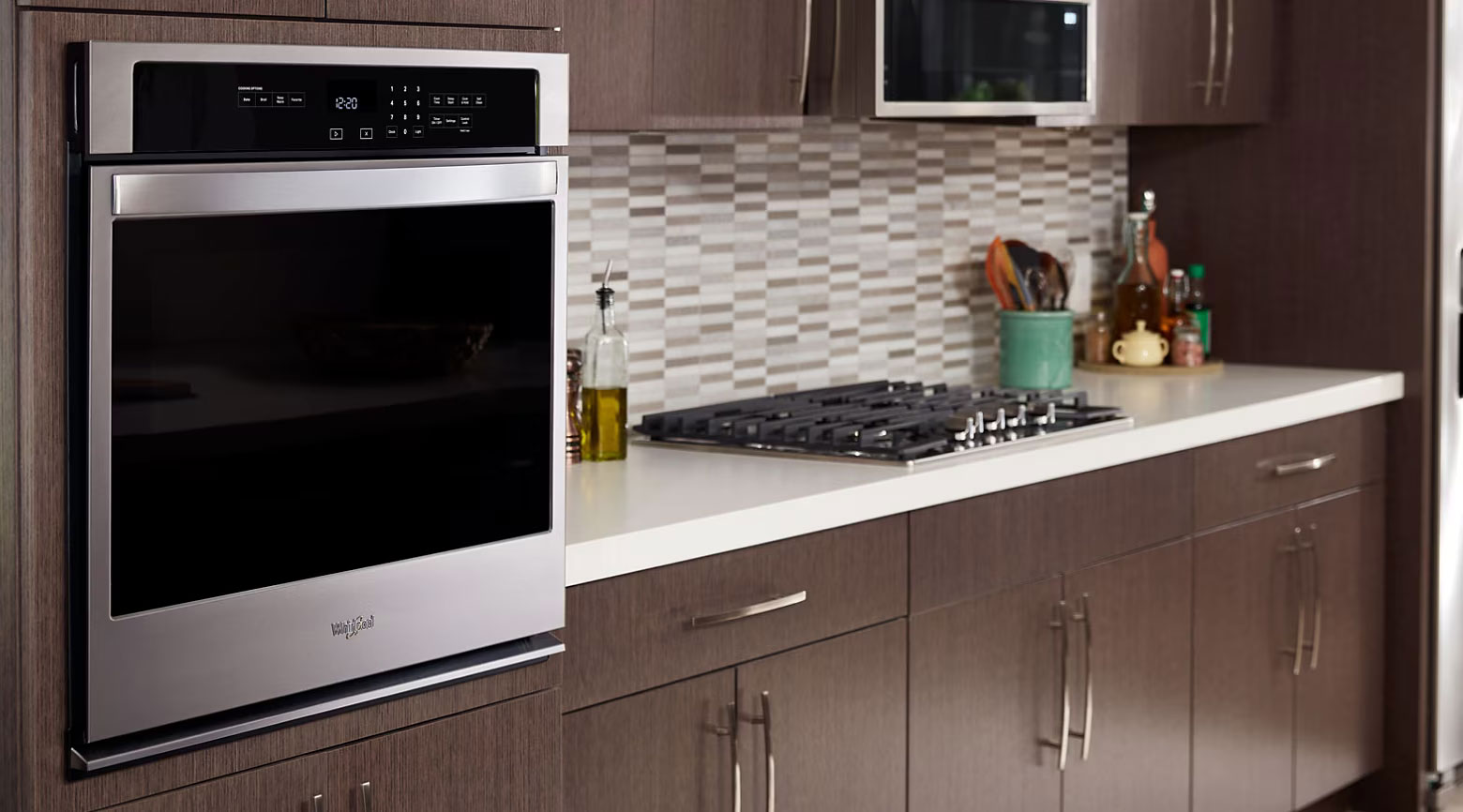 Whirlpool Combination Microwave Une cuisine avec un four mural Whirlpool et une table de cuisson Whirlpool à proximité. Sur les comptoirs, autour de la table de cuisson, sont posés une bouteille d'huile, un porte-ustensiles de cuisine et un plateau circulaire contenant des récipients, des bocaux et des bouteilles.