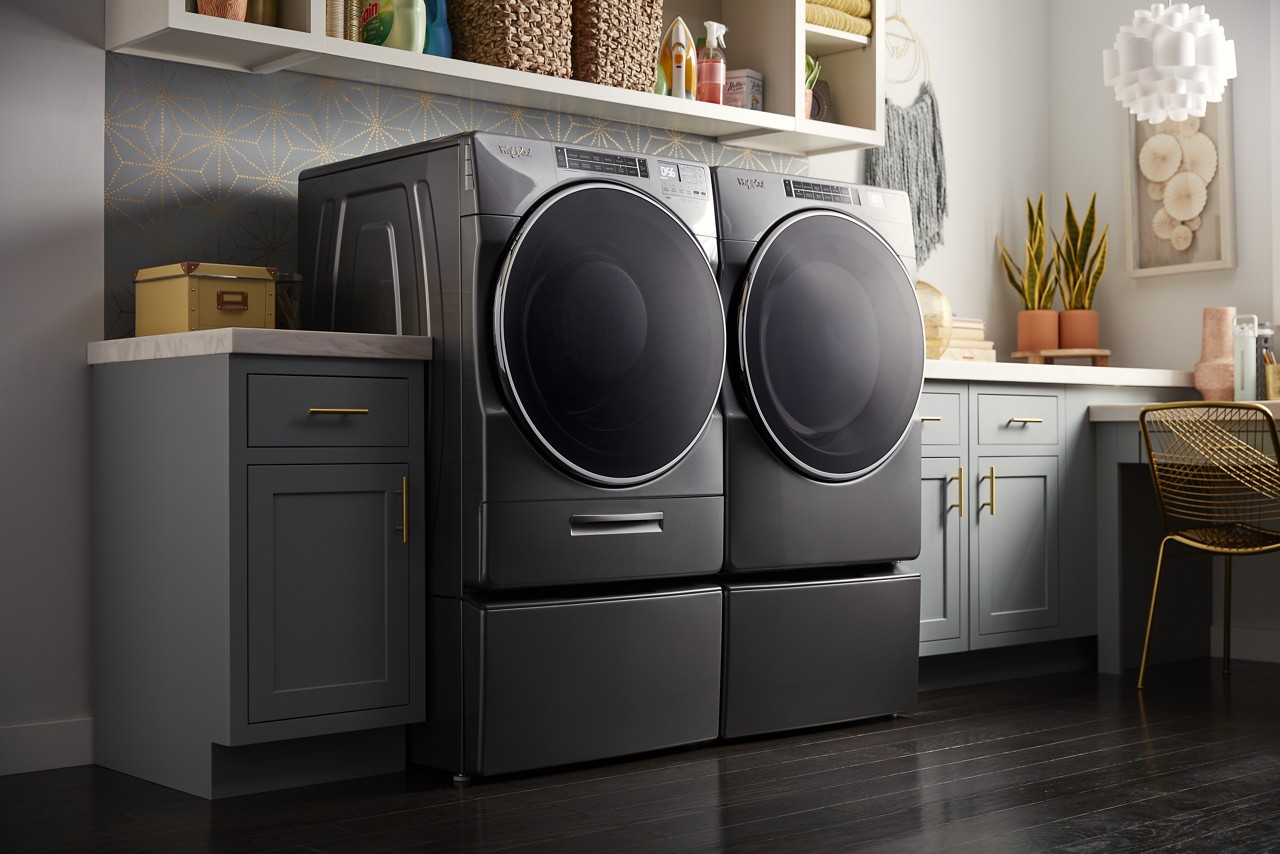 Un ensemble laveuse-sécheuse Whirlpool® dans une salle de lavage. Des paniers, des plantes en pot et une chaise glissée sous une table se trouvent à proximité.