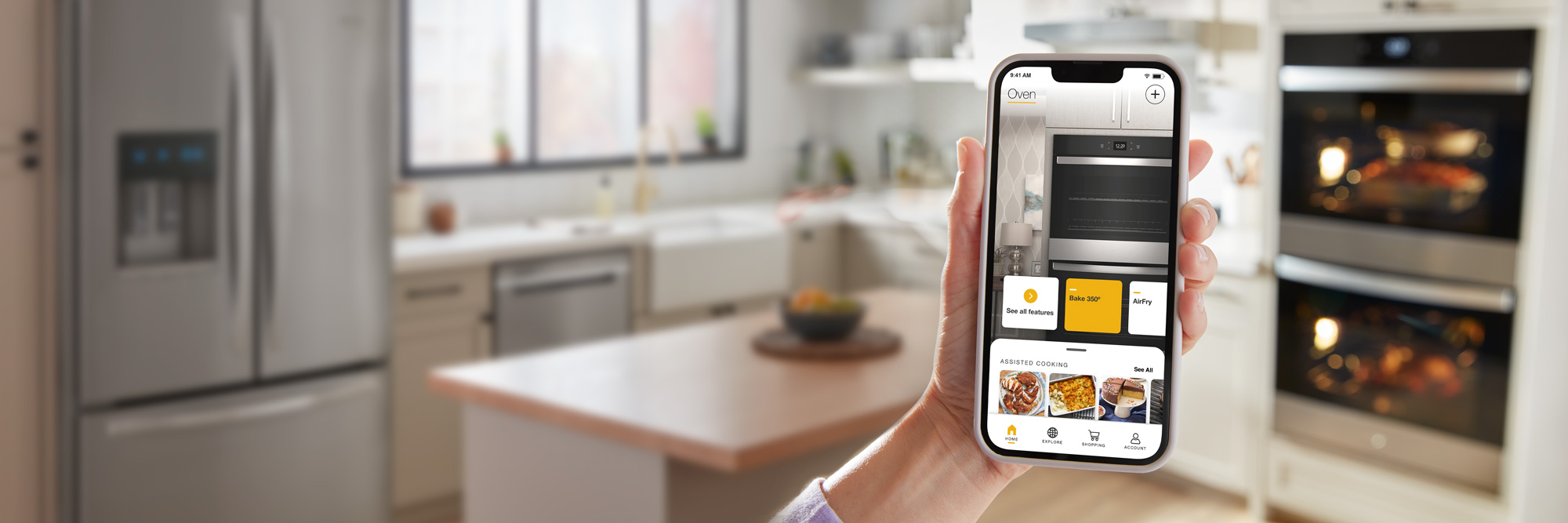A hand holds up a phone with the Whirlpool app open to the homepage with controls for the wall oven and assisted cooking features. In the background is a Whirlpool Double Wall Oven, each with the light on, a kitchen island and counters filled with various items