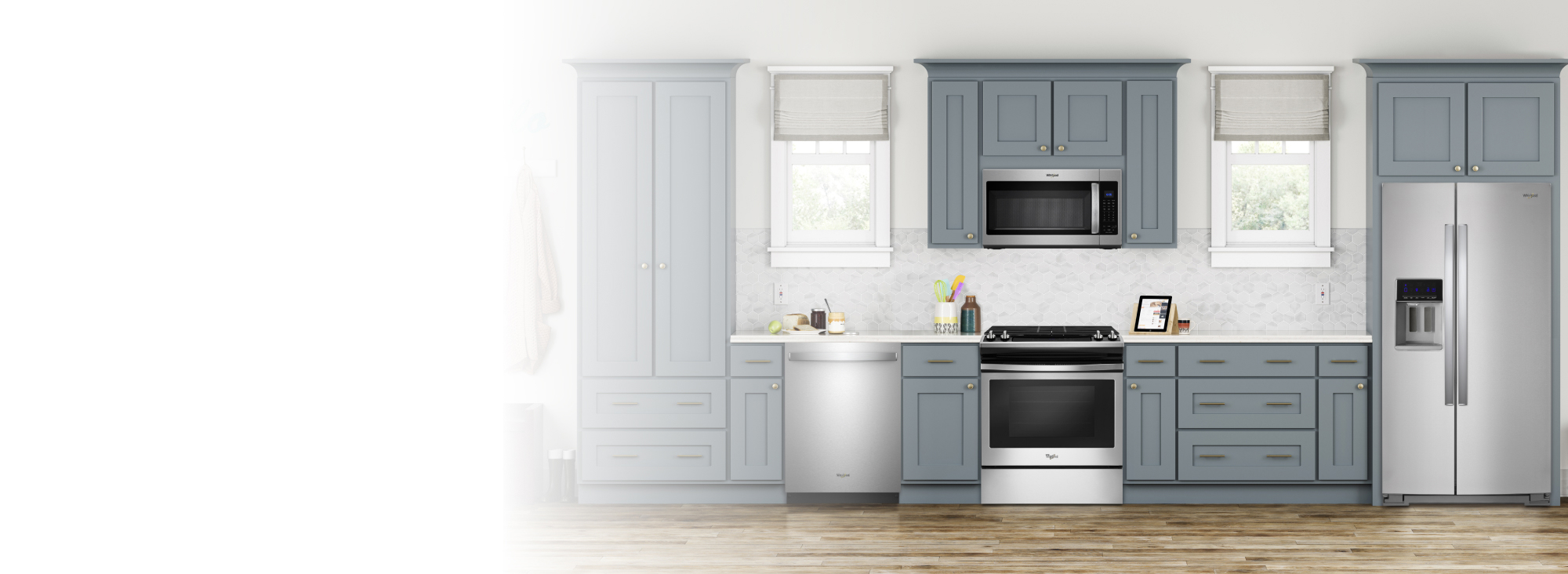 A kitchen with grey cabinets and two windows. There are several appliances, including a Whirlpool Range, Whirlpool Over-the-Range Microwave, Whirlpool Dishwasher and Whirlpool Refrigerator