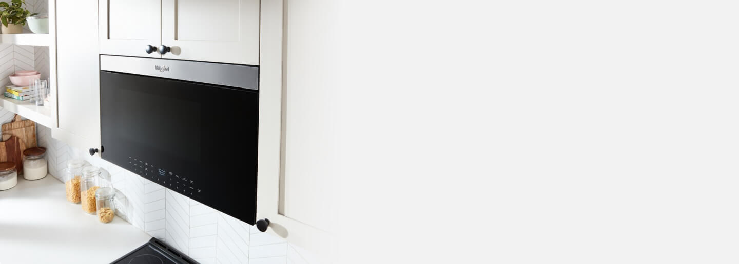 A flush Maytag Over-the-Range Microwave above a Maytag Cooktop surrounded by white cabinets. On the counters are jars of pasta and flour and cutting boards