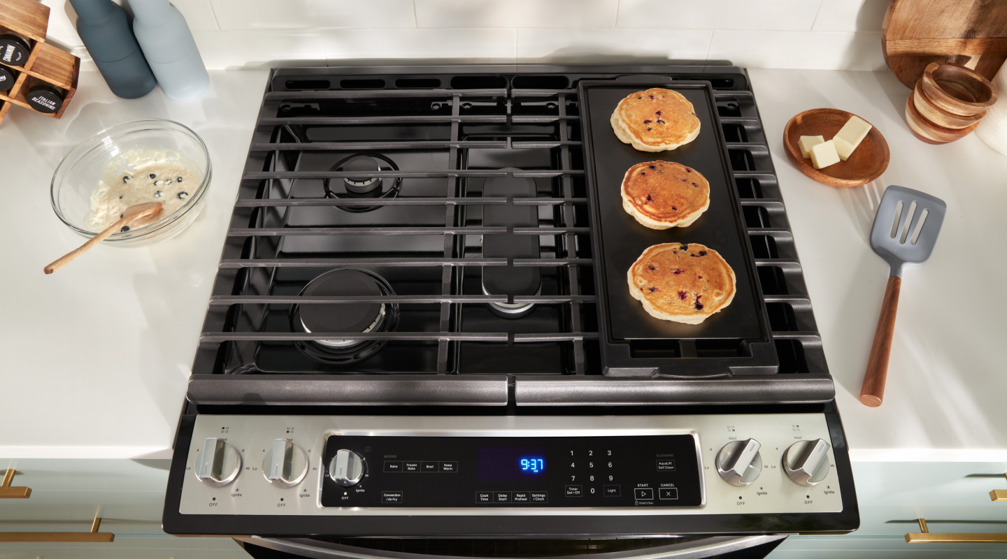 A finger presses a button on a Whirlpool Induction Range. There’s a pot with a liquid on one of the burners. On the counters surrounding the range are a wooden spoon on a dishcloth, containers and various items.