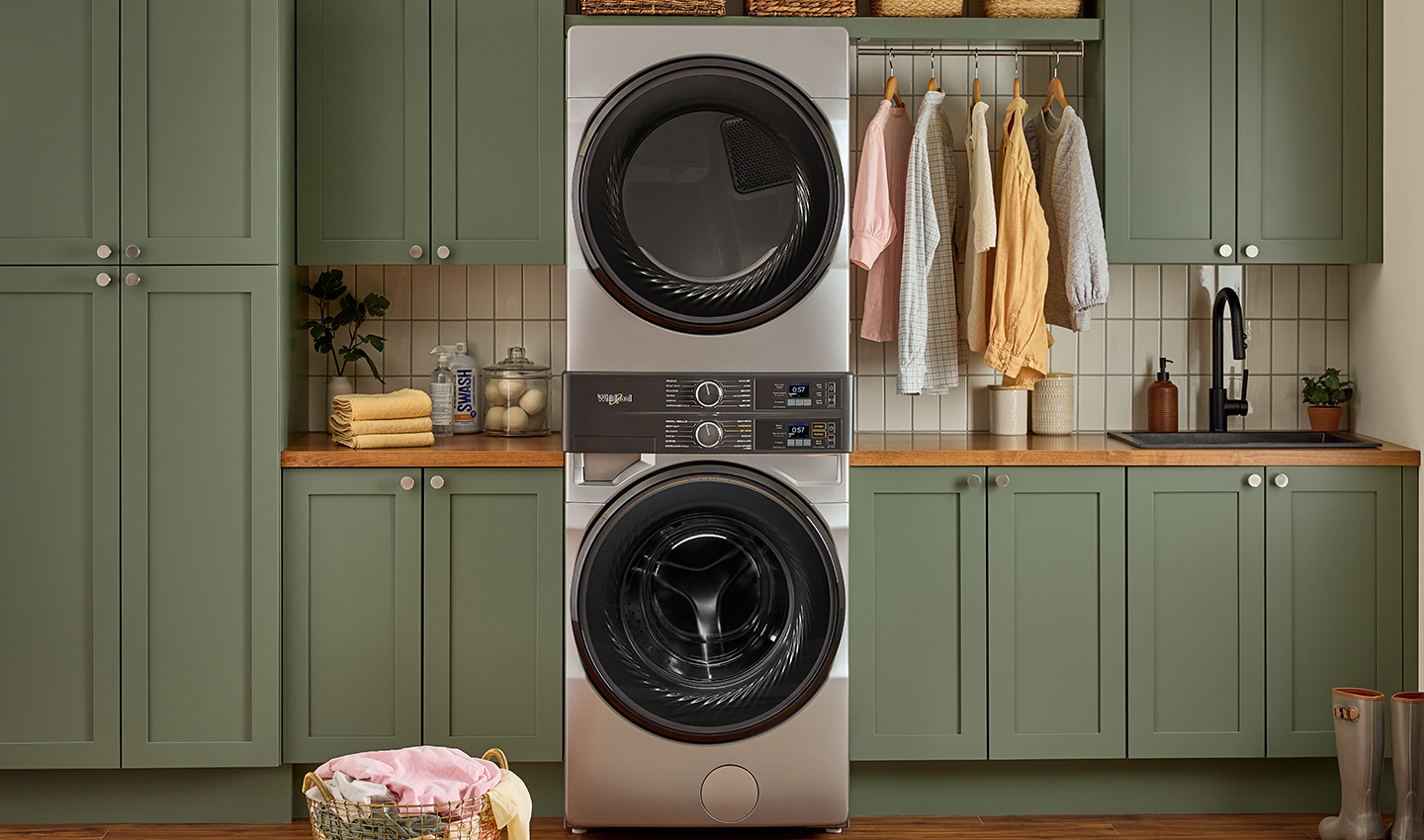 A Whirlpool laundry tower in a laundry room with green cabinets. In front is an overflowing laundry basket, and next to the appliances are hanging shirts