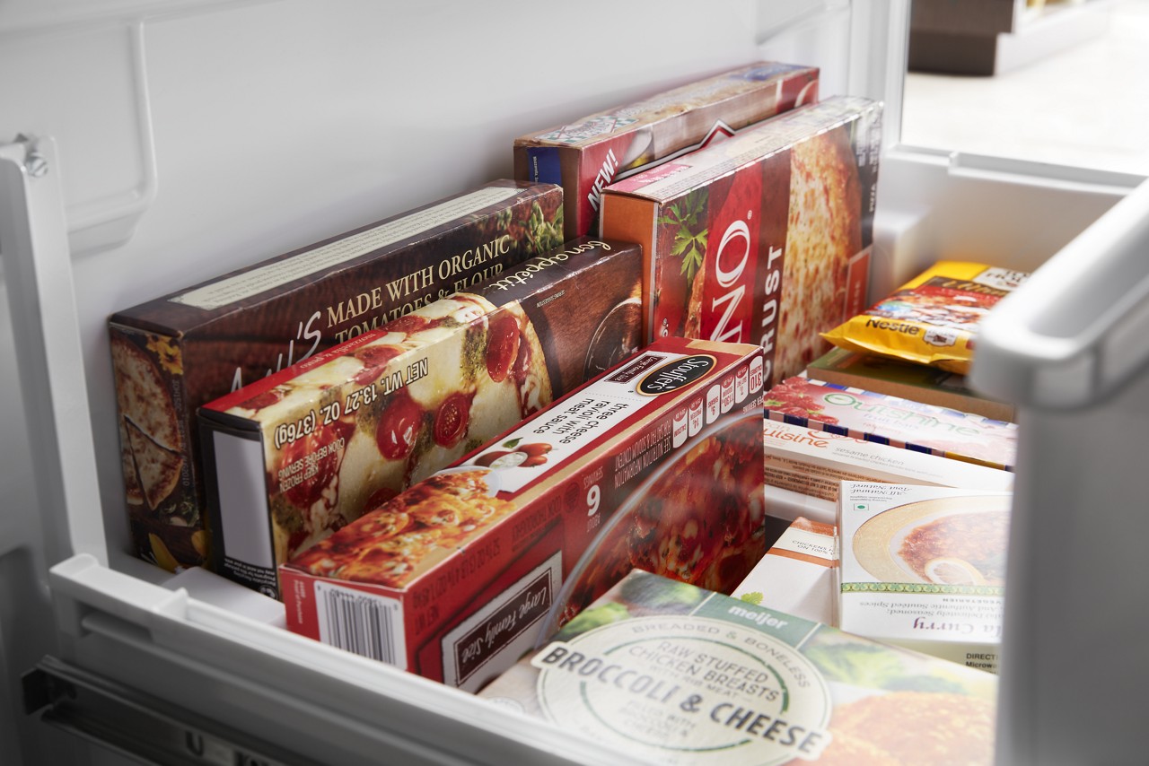 An open freezer drawer filled with frozen pizza and other frozen prepared foods. An open freezer drawer filled with frozen pizza and other frozen prepared foods.