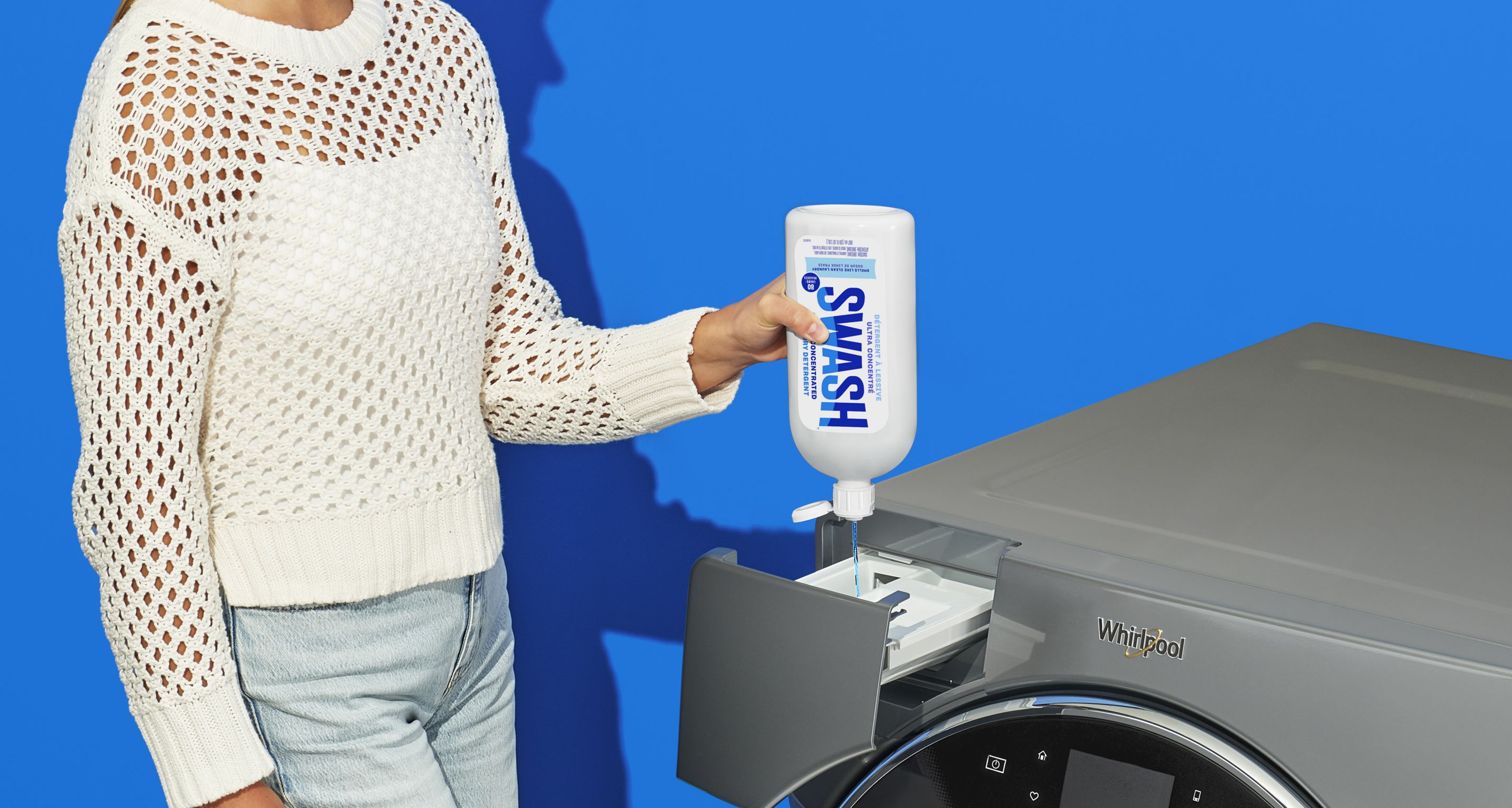  A woman squeezes detergent out of a bottle of Swash into the detergent dispenser of a Whirlpool Washing Machine in front of a blue background.
