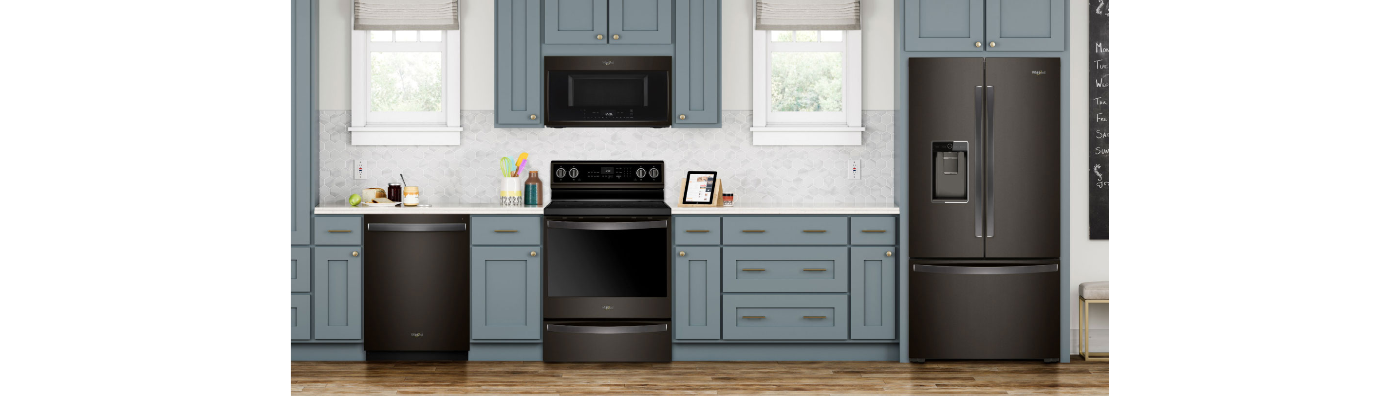 A kitchen with an island and three stools. On the island is a bowl of fruit on a placemat. The cupboards are grey and the Whirlpool Black Stainless Steel Appliances include a refrigerator, an oven with an over-the-range microwave and a dishwasher. On the counters are a kitchen utensil holder, cutting boards, a potted plant, salt and pepper shakers and more