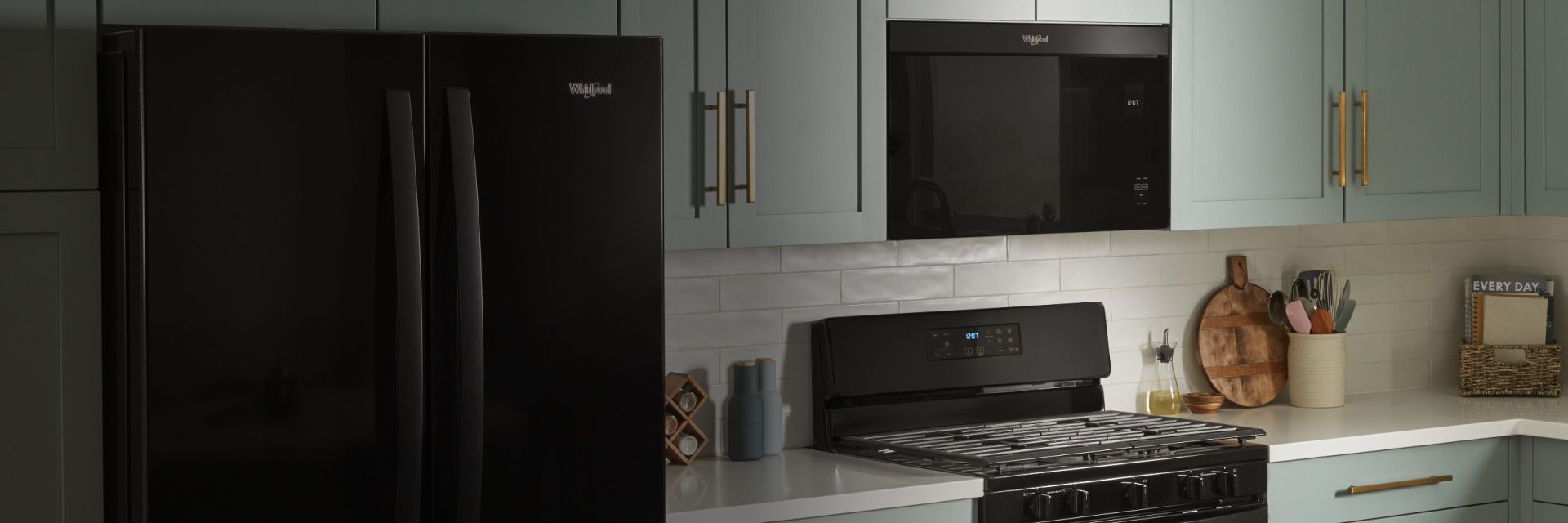 Une cuisine avec un réfrigérateur Whirlpool, un four à micro-ondes au-dessus de la cuisinière et une cuisinière. Sur les plans de travail se trouvent un porte-épices, un contenant d'huile, une planche à découper, un porte-ustensiles et un panier de livres de cuisine
