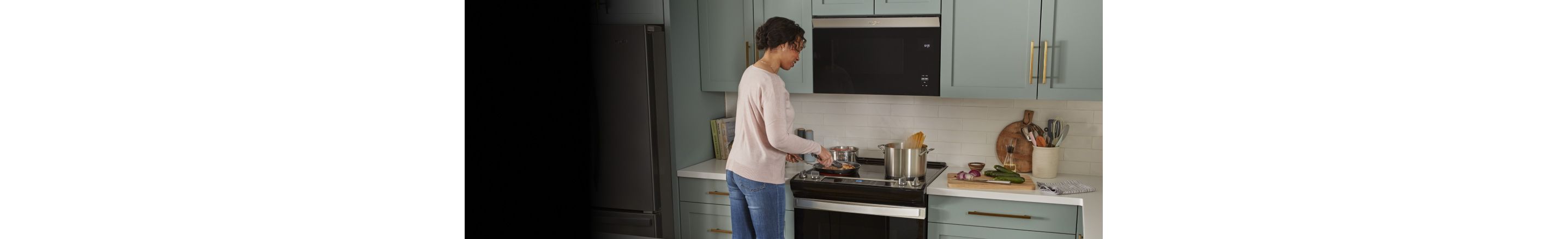 A woman cooks with a pan at a Whirlpool Oven below a flush over-the-range microwave. Also on the range are two pots, one of which has pasta cooking in boiling water. There's also a Whirlpool Refrigerator. On the counter are cookbooks, a cutting board with red onion, cucumbers and a knife, a dishcloth, a kitchen utensil holder and another cutting board leaning against the wall