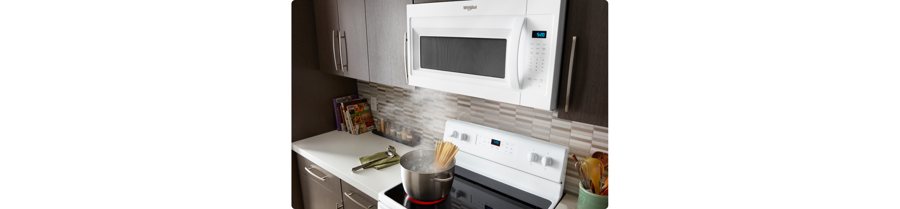 Des pâtes cuisent sur une cuisinière Whirlpool sous un four à micro-ondes à hotte intégrée Whirlpool. Sur les comptoirs se trouvent une louche posée sur un linge à vaisselle, des livres de cuisine et un porte-ustensiles