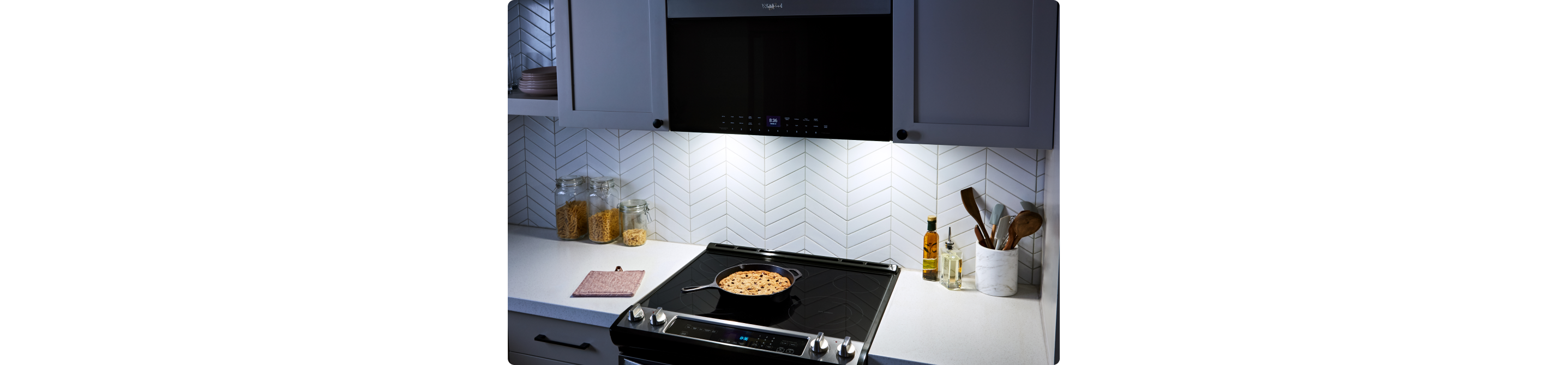 Un four à micro-ondes à hotte intégrée Whirlpool au un design affleurant. Sa lumière éclaire la table de cuisson et une casserole en dessous. Sur le comptoir se trouvent un porte-ustensiles, des contenants d'huile et des bocaux de pâtes
