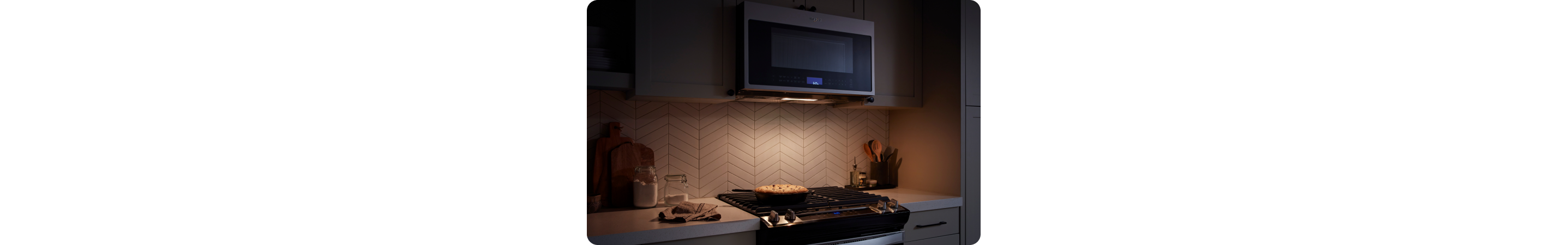 Dans une cuisine sombre se trouve un four à micro-ondes Whirlpool au-dessus d'une table de cuisson Whirlpool. La lumière de la table de cuisson est allumée et on voit une poêle en train de cuire des aliments. Sur les comptoirs se trouvent des planches à découper, un porte-ustensiles et autres objets