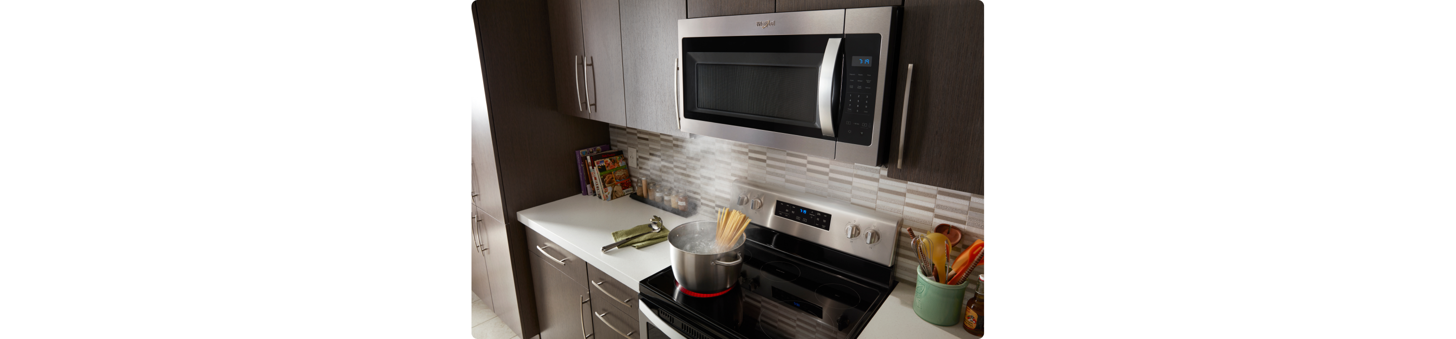 Une cuisine avec un four à micro-ondes à hotte intégrée Whirlpool au-dessus d'une cuisinière Whirlpool. Sur la table de cuisson à induction, des pâtes cuisent dans une casserole d'eau. Sur les comptoirs se trouvent un porte-ustensiles, un ustensile sur un chiffon et des livres de cuisine