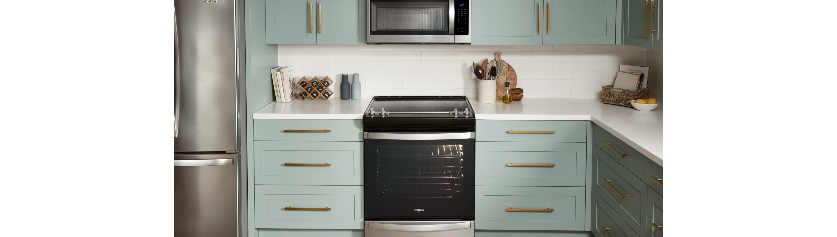 A kitchen with a Whirlpool Refrigerator and a Whirlpool Range with an Over-the-Range Microwave. On the counters are cookbooks, a spice rack, a full kitchen utensil holder, a cutting board and more