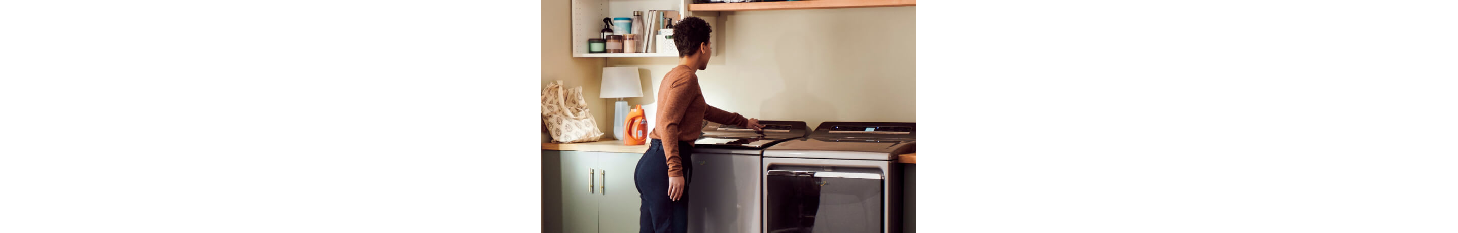 Une femme place des vêtements dans une laveuse  à chargement par le haut Whirlpool. À côté de la laveuse se trouve une sécheuse Whirlpool. Sur les comptoirs sont posés des bouteilles de détergent et d'autres objets