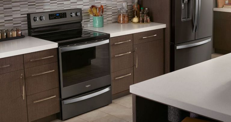 A kitchen with a Whirlpool stove and a Whirlpool refrigerator. The kitchen features brown cabinets and drawers. On the counters are containers of herbs and spices, a container with kitchen utensils and various bottles and jars.