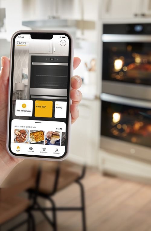 A hand holds up a phone with the Whirlpool app open to the homepage with controls for the wall oven and assisted cooking features. In the background is a Whirlpool Double Wall Oven, each with the light on, a kitchen island and counters filled with various items