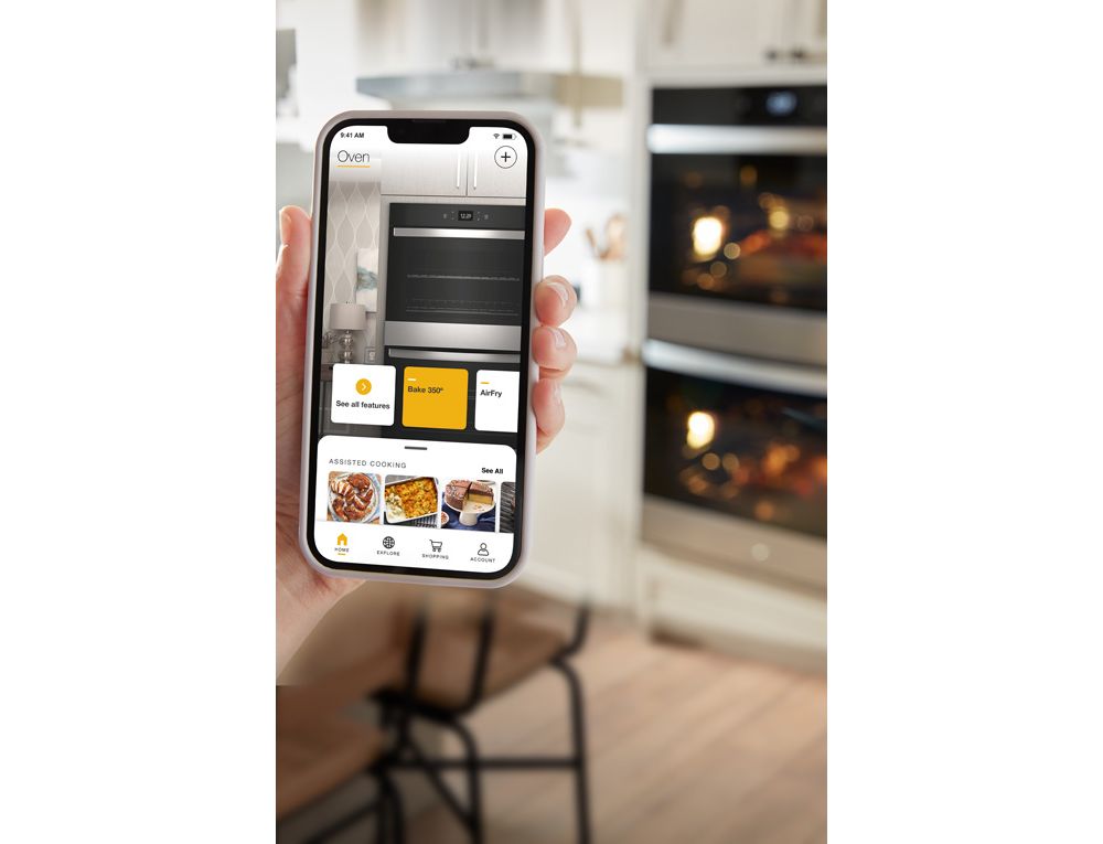 Une main tient un téléphone où l'application Whirlpool est ouverte sur la page d'accueil avec les commandes pour le four mural et les fonctions de cuisson assistée. En arrière-plan, on voit un four mural double Whirlpool avec la lumière allumée, un îlot de cuisine et des comptoirs remplis de différents objets